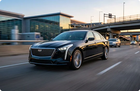 A black Cadillac Sedan in fast motion on a roadway with glass building and an overhead roadway in background.