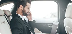 A businessman in formal attire speaks on the phone while working on his laptop in a luxury car.