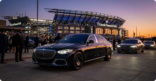 A black Mercedes-Benz Maybach sedan is parked in a paved lot in front of Lincoln Financial Field at dusk. The stadium is illuminated with blue lights against a purple and orange sunset sky, with several people and other vehicles in the background.