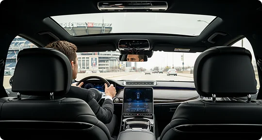 A professional chauffeur in a suit drives a luxury car with a panoramic sunroof, showing a high-tech digital dashboard and a view of a stadium through the windshield.