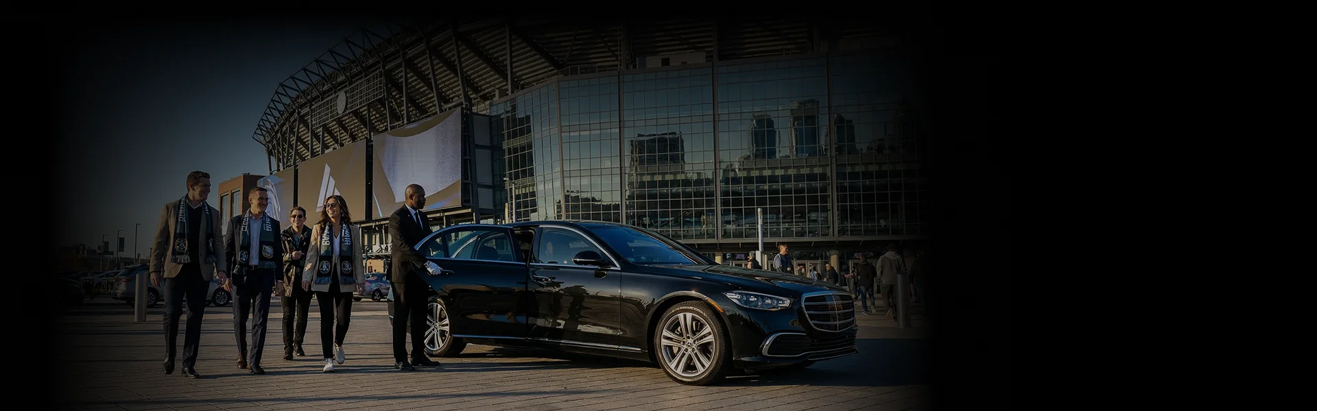 A professional chauffeur in a suit and white gloves opens the rear door of a black luxury sedan for a group of four smiling sports fans wearing team scarves, with a large modern stadium in the background.
